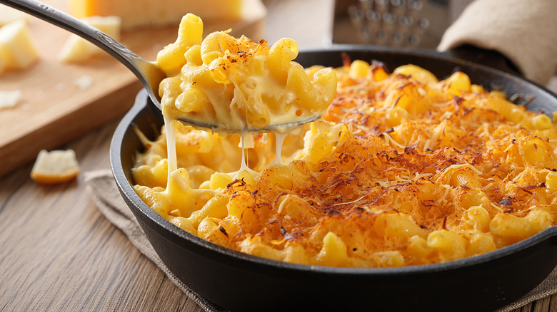 A spoonful of crispy-topped mac and cheese pulling away from a cast iron pan full of the dish