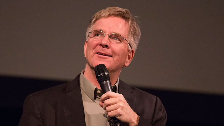 Rick Steves wearing a brown suit and holding a microphone