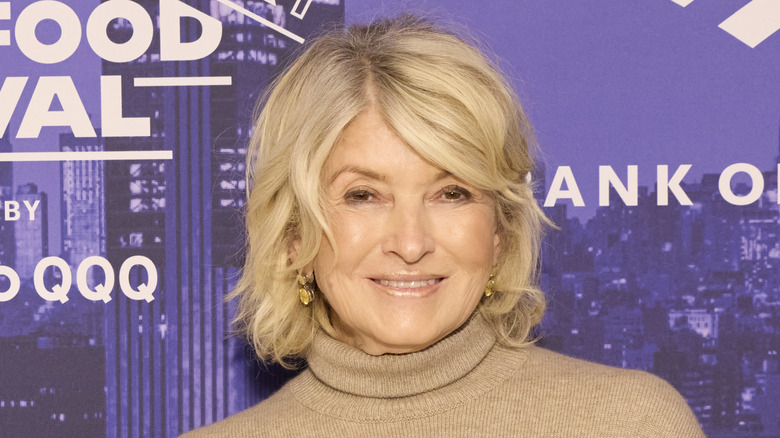 Martha Stewart smiling in front of purple marketing wearing brown turtleneck sweater