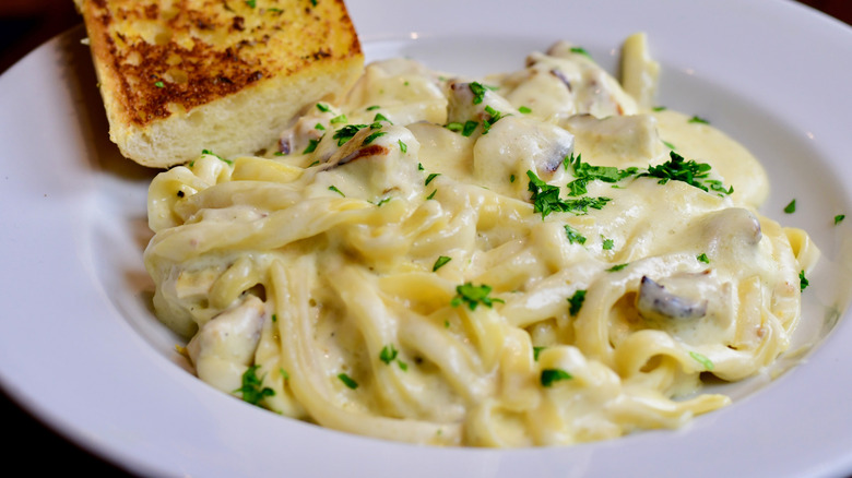White dish of fettuccine Alfredo pasta with parsley and a slice of garlic bread