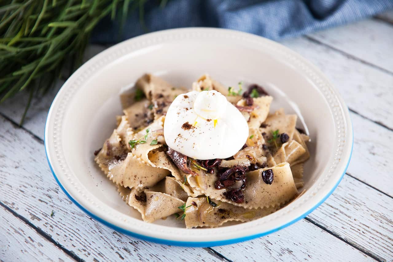 A white bowl with a blue rim sits on a white-washed wooden surface. The bowl holds ribbons of pasta, topped with a round of mozzarella.