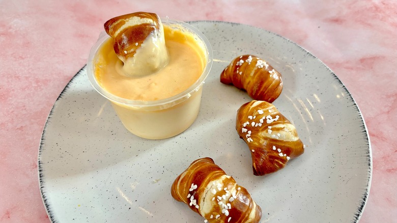 White plate with three pretzel bites, one dipped in cup of cheese, all on pink marble background