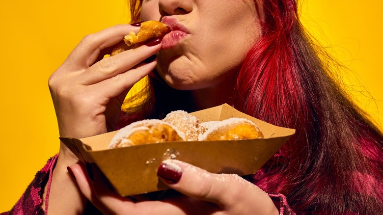 Cropped image of woman enjoying delicious mini donuts with powdered sugar, eating food with pleasure against bright yellow background. Concept of street food, winter fairs, pop art, holiday snacks
