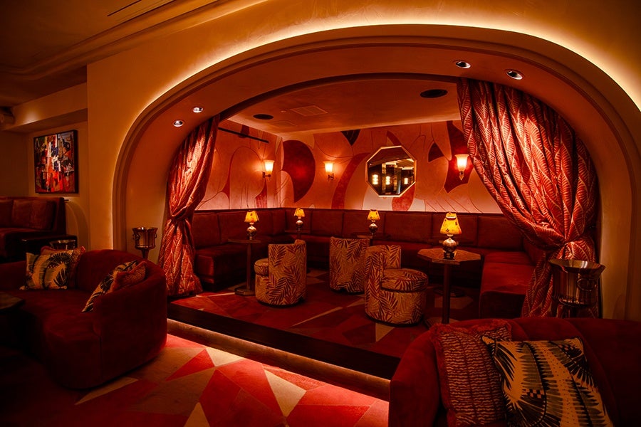 A lounge area in an upscale cocktail bar, set off by heavy red curtains, with red velvet couches.