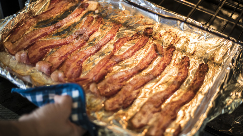 A person pulling a baking sheet with bacon on it out of the oven