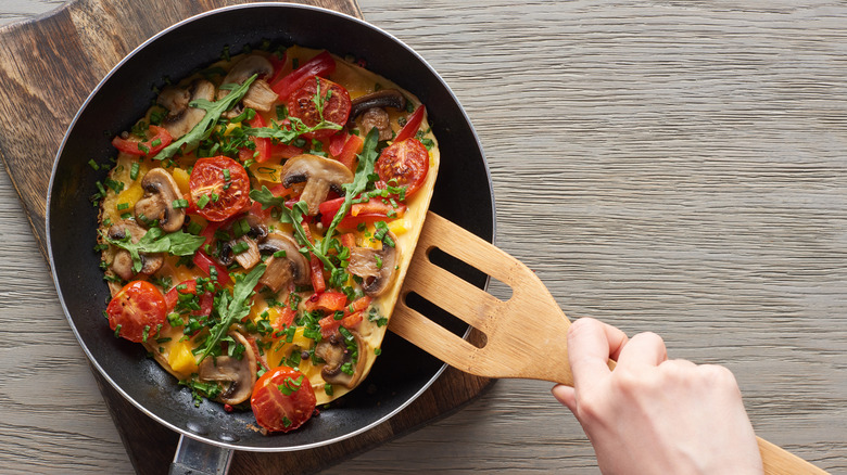 Cropped view of hands cooking omelet with mushrooms, tomatoes, and greens on frying pan with wooden slotted spatula