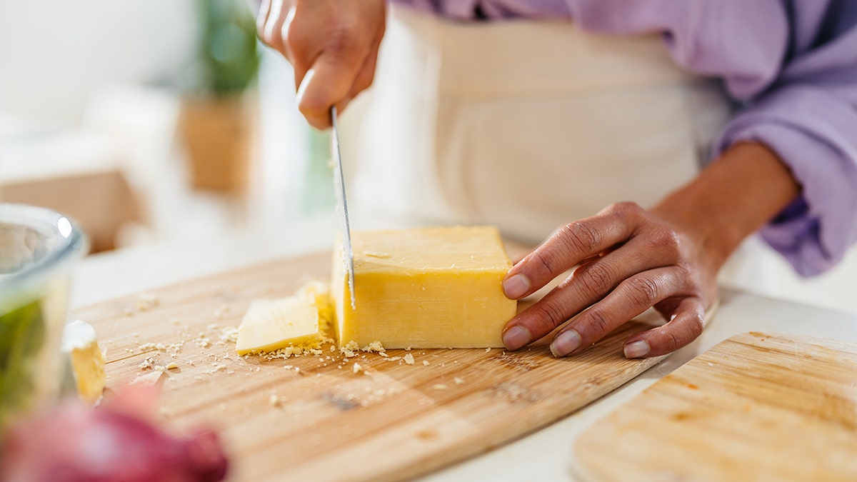 Hands slicing a block of cheese with a knife on a wooden cutting board in a kitchen.