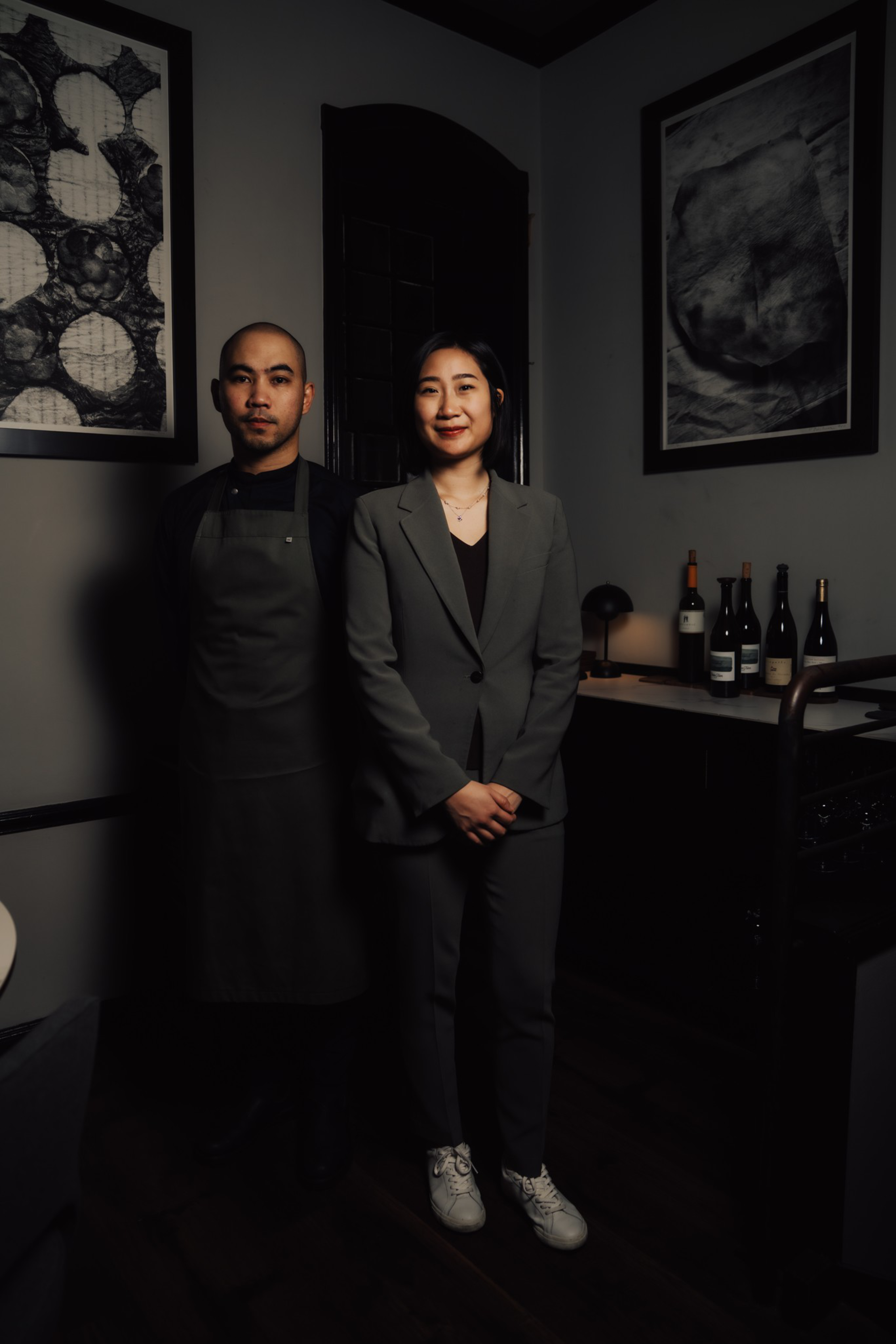 A man in a dark apron and a woman in a gray suit stand side by side in a dimly lit room with framed black-and-white artwork and bottles on a counter behind them.