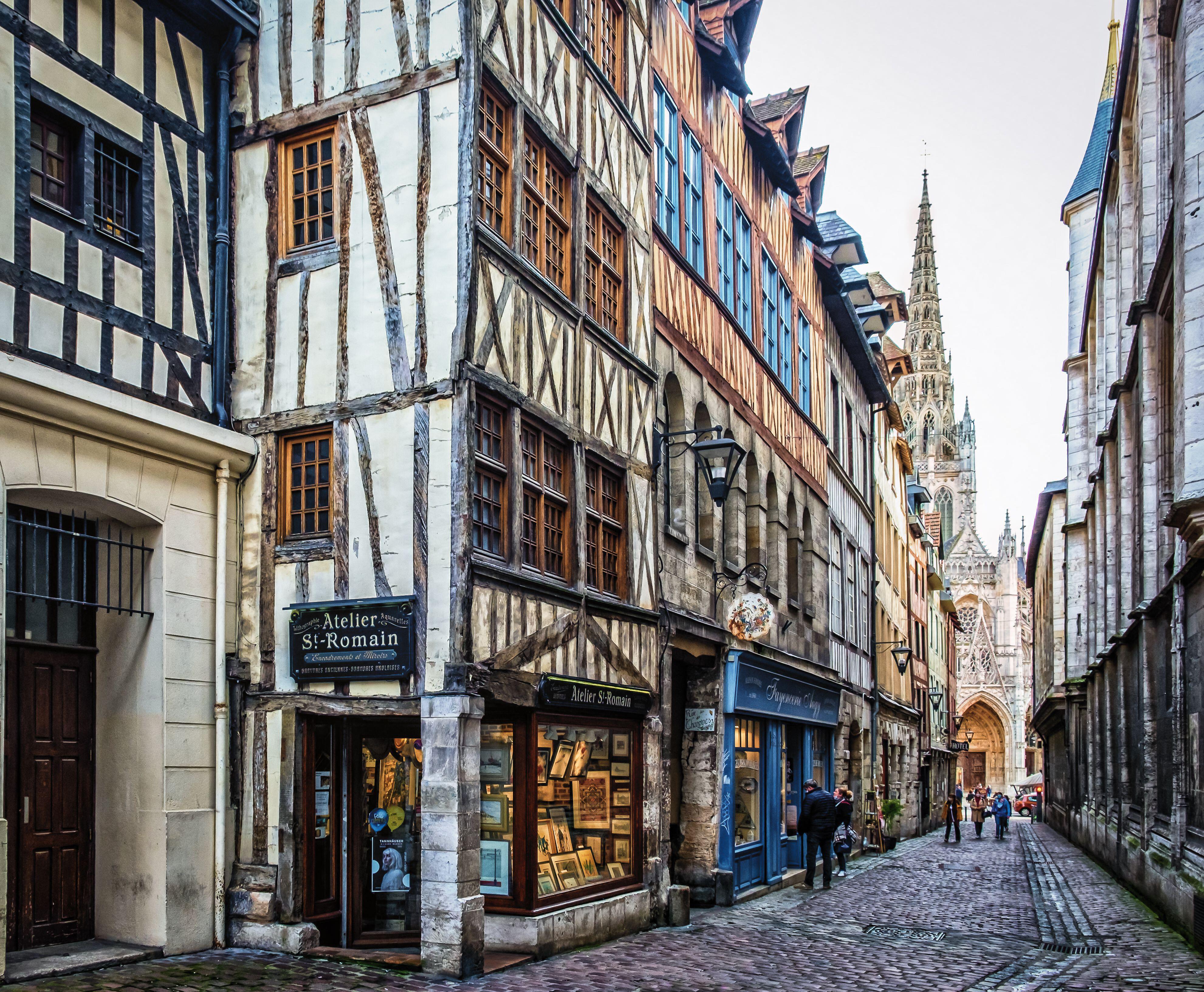 View of Rue Saint-Romain, a cobblestoned street in Rouen, France, lined with medieval half-timbered houses, leading to a gothic cathedral spire in the background.