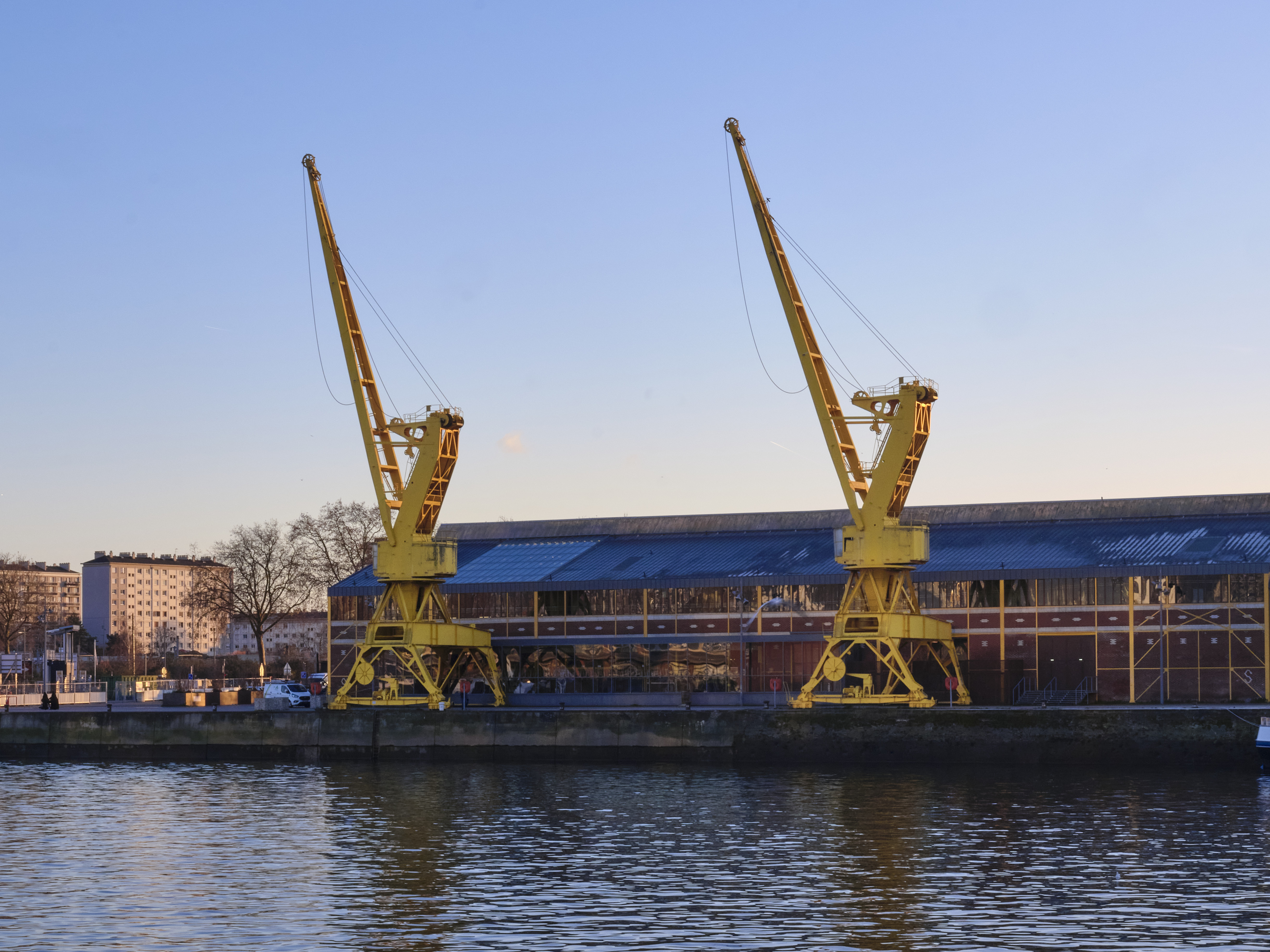 Two yellow cranes stand by a building with a blue roof next to a body of water.
