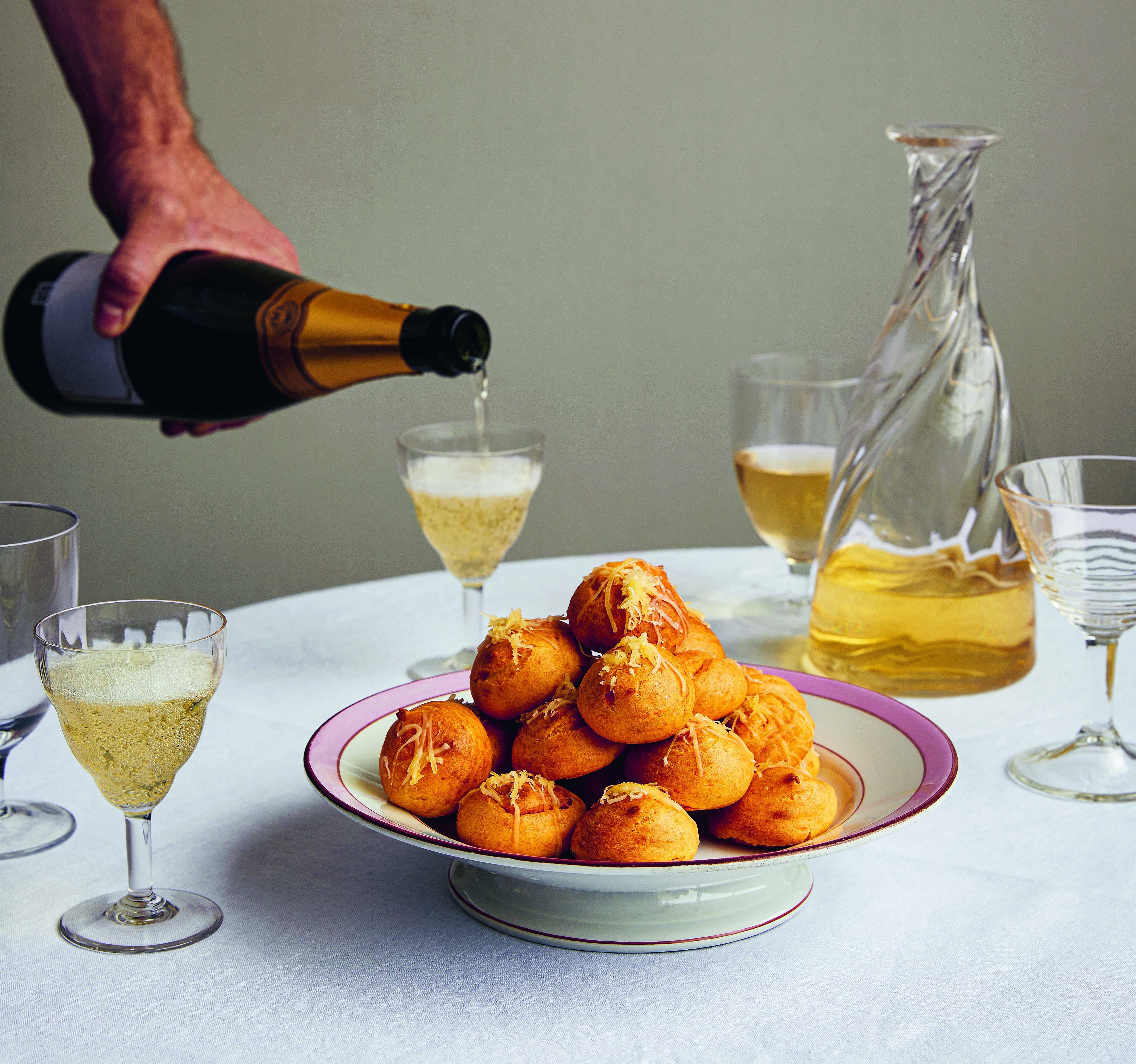 A person pouring champagne into a glass next to a plate of Comte Gougères.