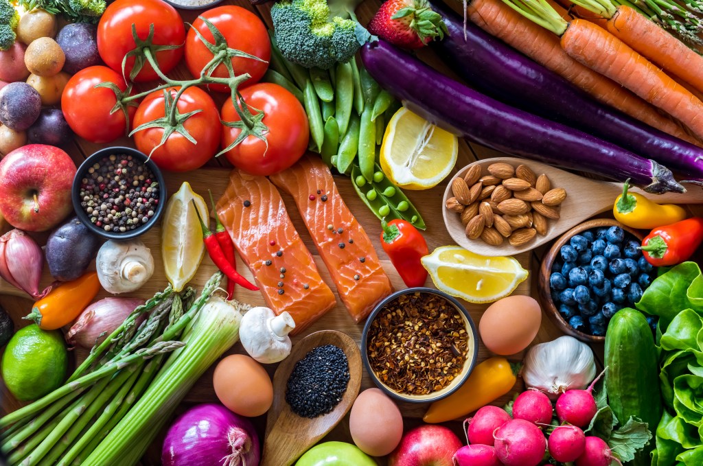 Flat-lay arrangement of fresh produce including fruits, vegetables, eggs, nuts, spices, and salmon fillets.