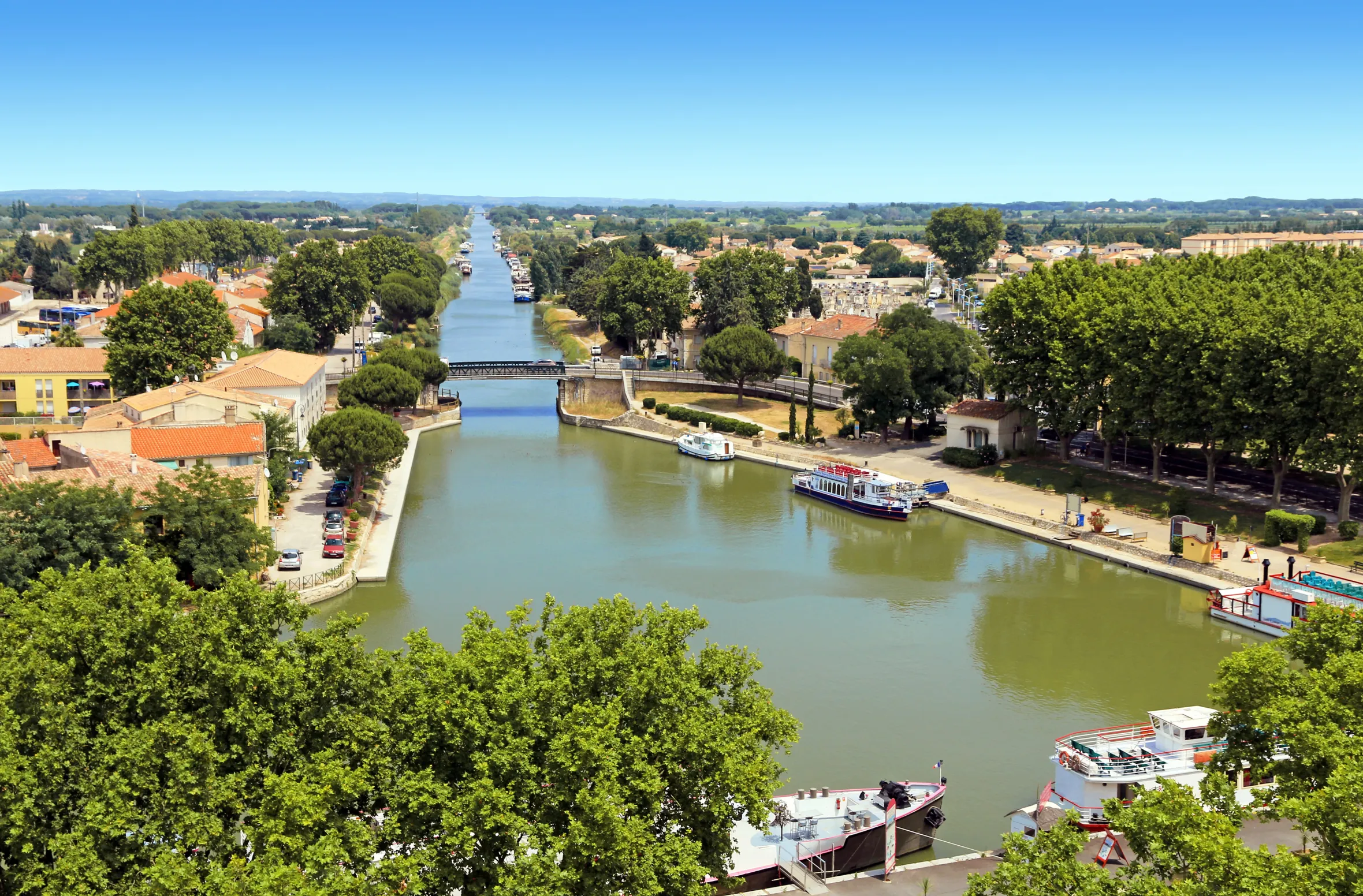 Aerial view of the Rhône canal in Sète, connecting Etang de Thau to the Rhône at Beaucaire, with boats docked along the banks and buildings surrounded by trees.