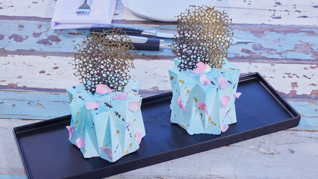 Two small cakes shaped with origami folds on the icing sides. Topped with a webbing of edible tuile and garnished with pink wafer peteas.