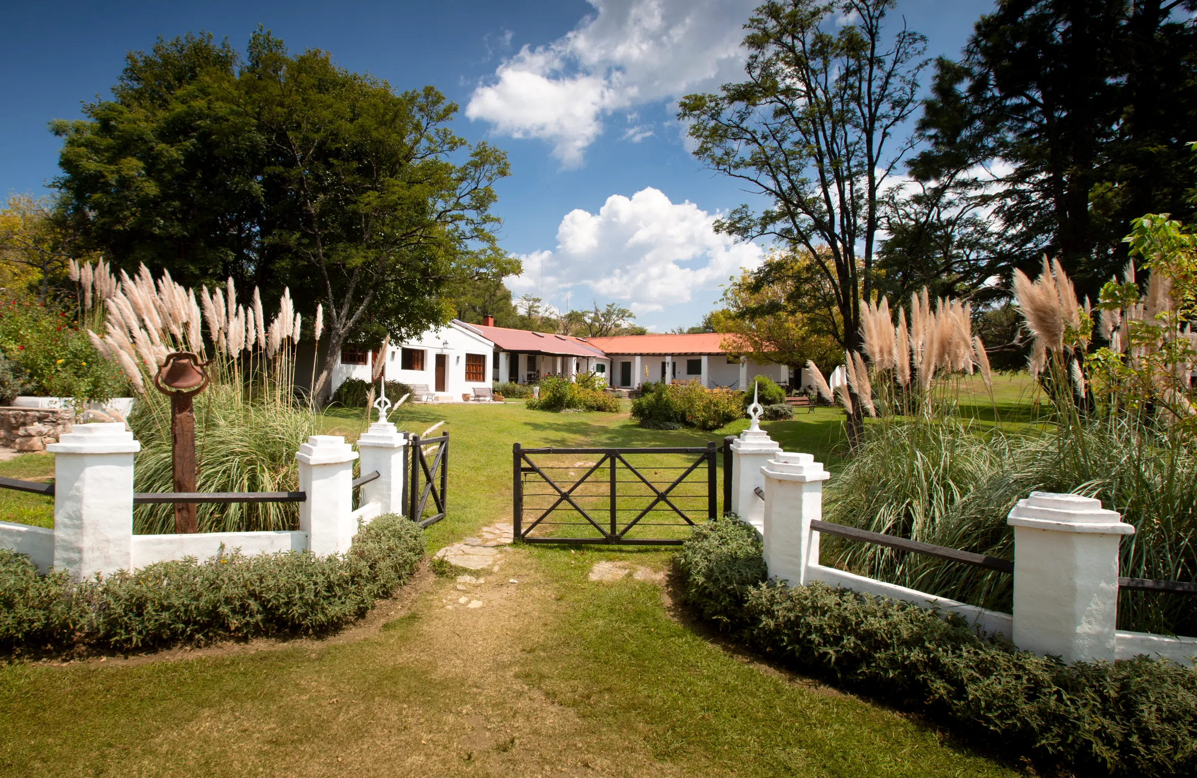 A white house with a red roof seen through a black gate, surrounded by trees and tall grass, under a blue sky with white clouds.