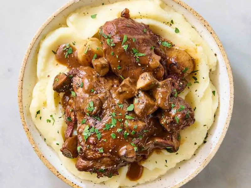 Tender chicken pieces with mushrooms in a rich gravy served over creamy mashed potatoes, garnished with parsley.