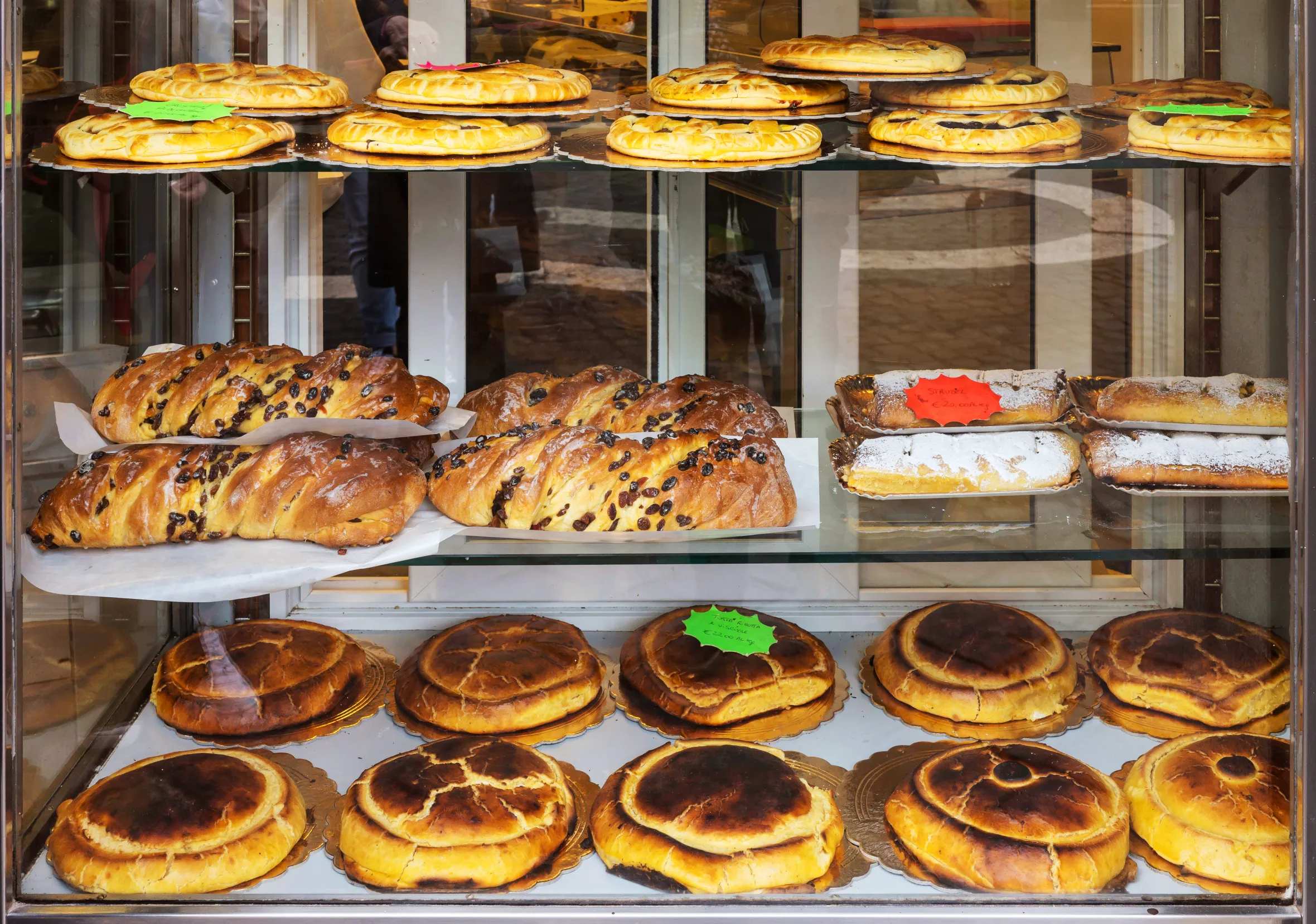 Pastries, including Crostata, strudel, Pizza Ebraica, and Torta Ricotta e Visciole, displayed in a shop window.