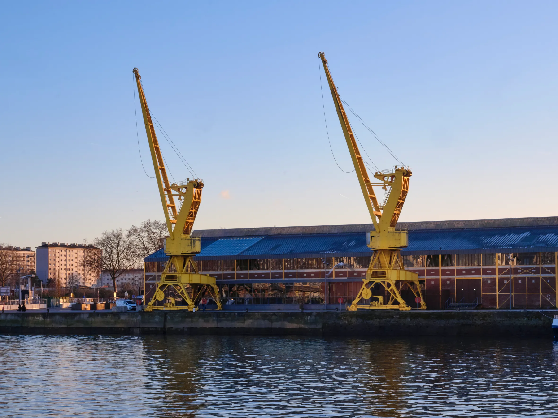 Two yellow cranes stand by a building with a blue roof next to a body of water.