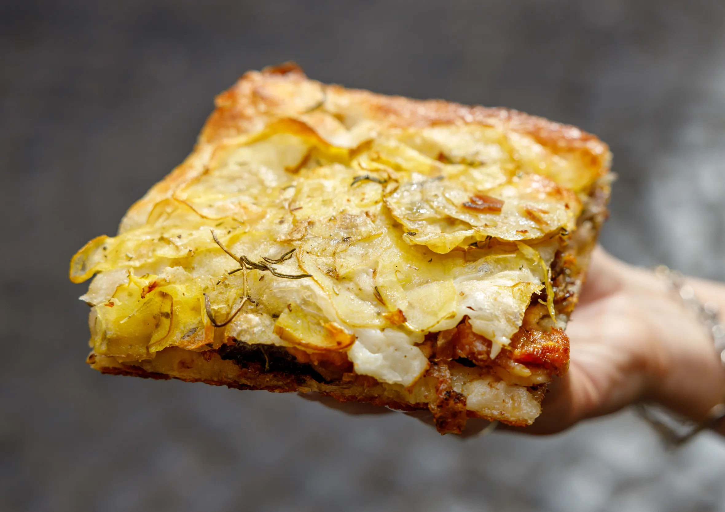 A hand holds a square slice of potato focaccia from Roscioli's in Rome, with a blurry background.