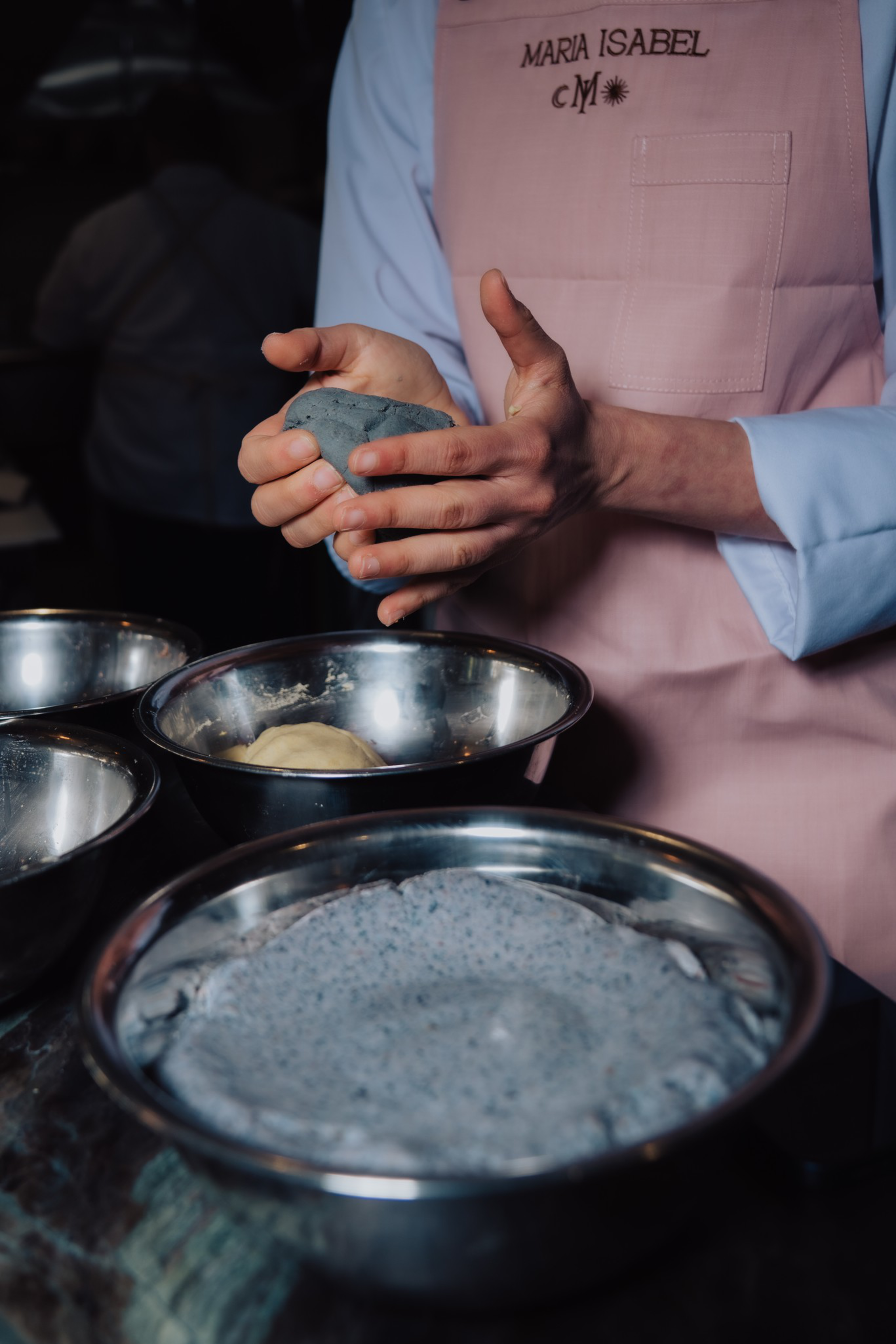 Hands wearing a pink apron shape blue dough above metal bowls containing blue and beige dough on a dark countertop.