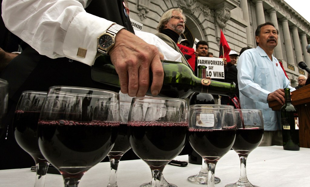 A man in a white shirt and watch pouring Gallo wine into glasses, with UFW President Arturo Rodriguez speaking in the background.