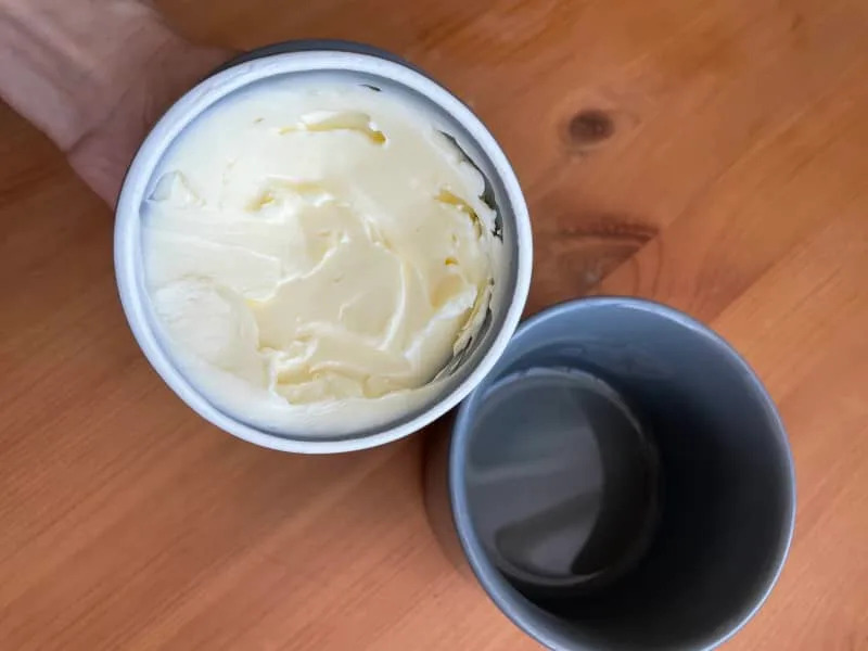 Hand holding a container of creamy butter next to an empty gray cup on a wooden table.