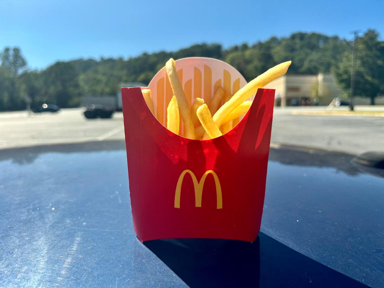 A red McDonald's container filled with French fries sits on a car hood in a sunny parking lot, with trees and buildings blurred in the background.