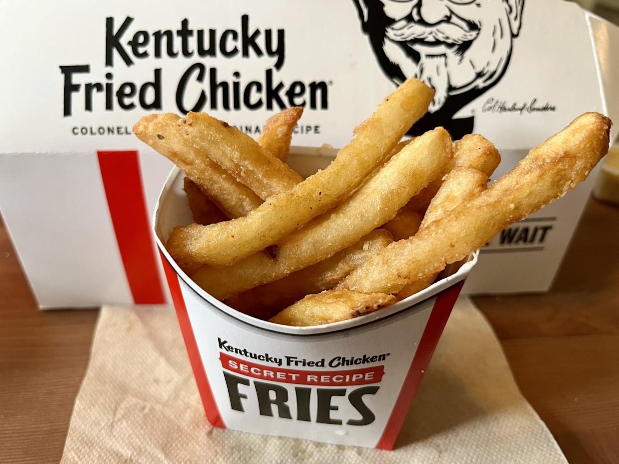 Close-up of a container of KFC Secret Recipe Fries on a napkin, with a KFC takeout box in the background featuring the brand logo and Colonel Sanders’ face.