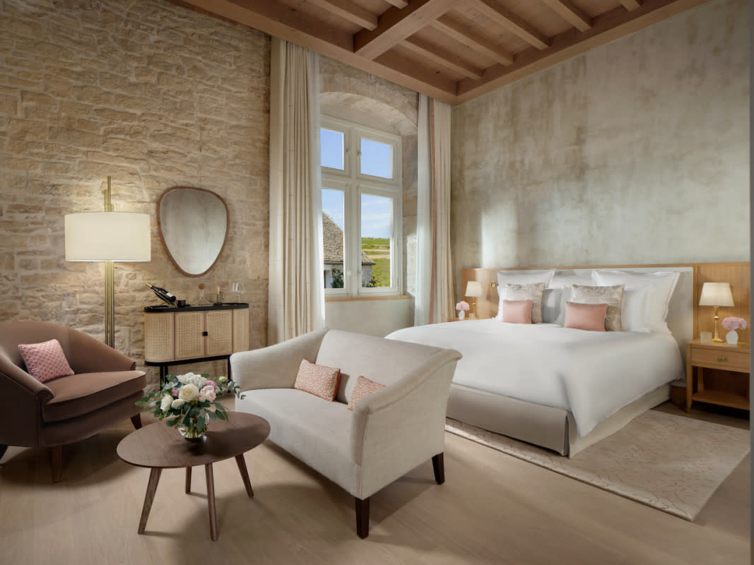 Hotel & Resort PhotographyThe interior of a luxury bedroom suite featuring a mix of rustic and modern design&period; One wall is made of exposed natural stone&comma; while the other is a soft&comma; textured grey&period; The room includes a large bed with white linens and pink accent pillows&comma; a cream-colored loveseat&comma; and a mid-century modern lounge chair&period; Large windows with white drapes look out onto a sunlit vineyard&comma; and a vaulted wooden ceiling spans the top of the room&period;