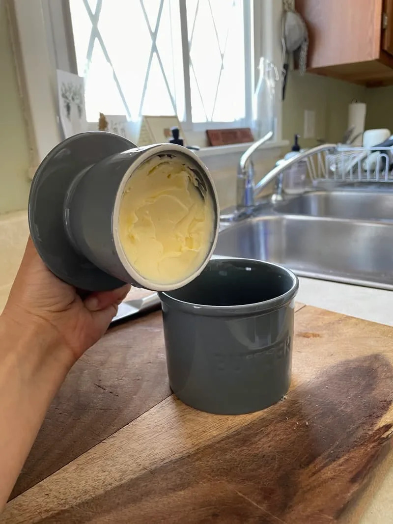 Hand holding a butter bell with fresh butter over a wooden countertop in a kitchen with a sink and window.