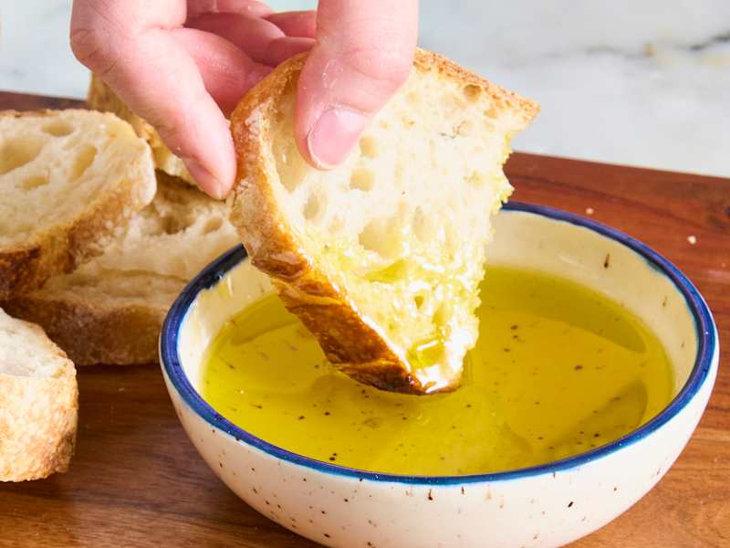 A hand dipping a piece of crusty bread into a small bowl of olive oil on a wooden surface.