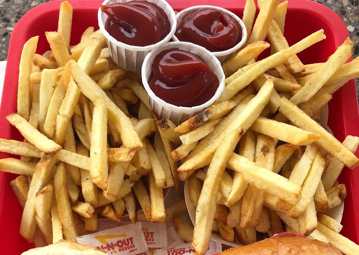 A red tray filled with a large serving of French fries, three paper cups of ketchup, and packets of In-N-Out Burger sauce partially visible at the bottom.
