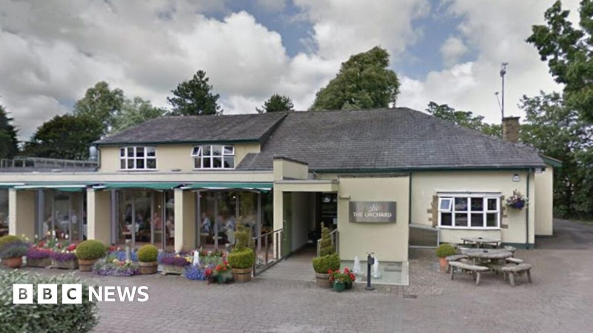 Street view image of the Italian Orchard in Broughton. A long cream rendered building with a grey roof and a white bow window to the right. To the left are three large glass window areas with many flowers and pots outside