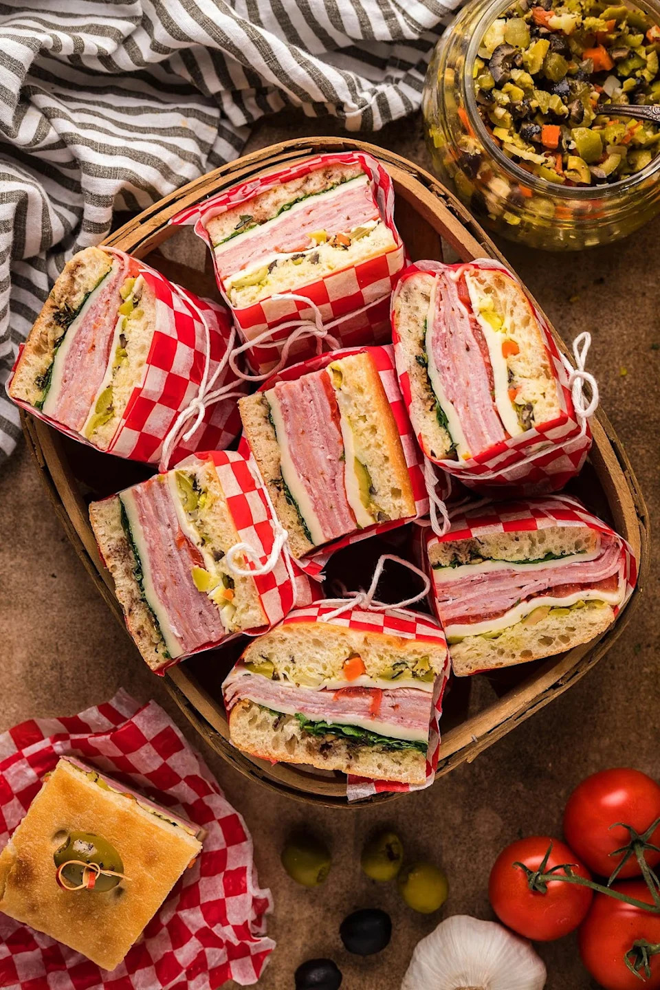 Pressed Italian picnic sandwiches in a picnic basket with a sandwich next to it on checkered paper and other ingredients on the table.