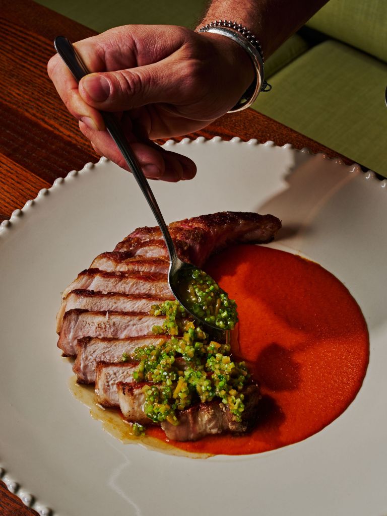 Chef spooning herb salsa verde over sliced grilled pork chop on red pepper purée at an upscale restaurant.