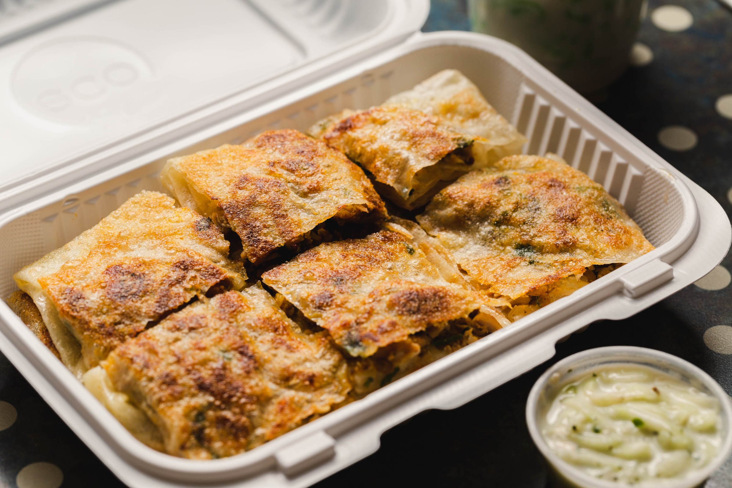 A rectangular takeout container filled with cut-up pieces of keema palata and a small container of dip nearby.