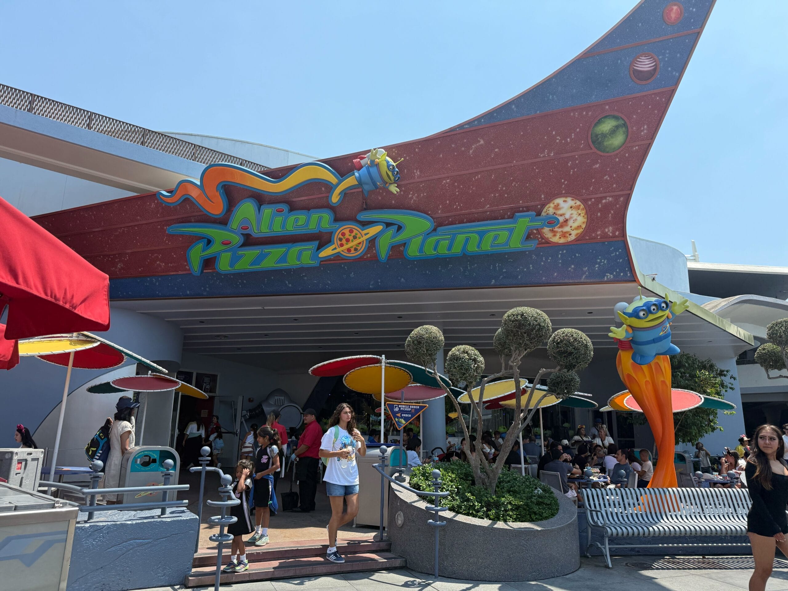 People walk past the colorful Alien Pizza Planet restaurant with a space-themed sign and decorations outside.