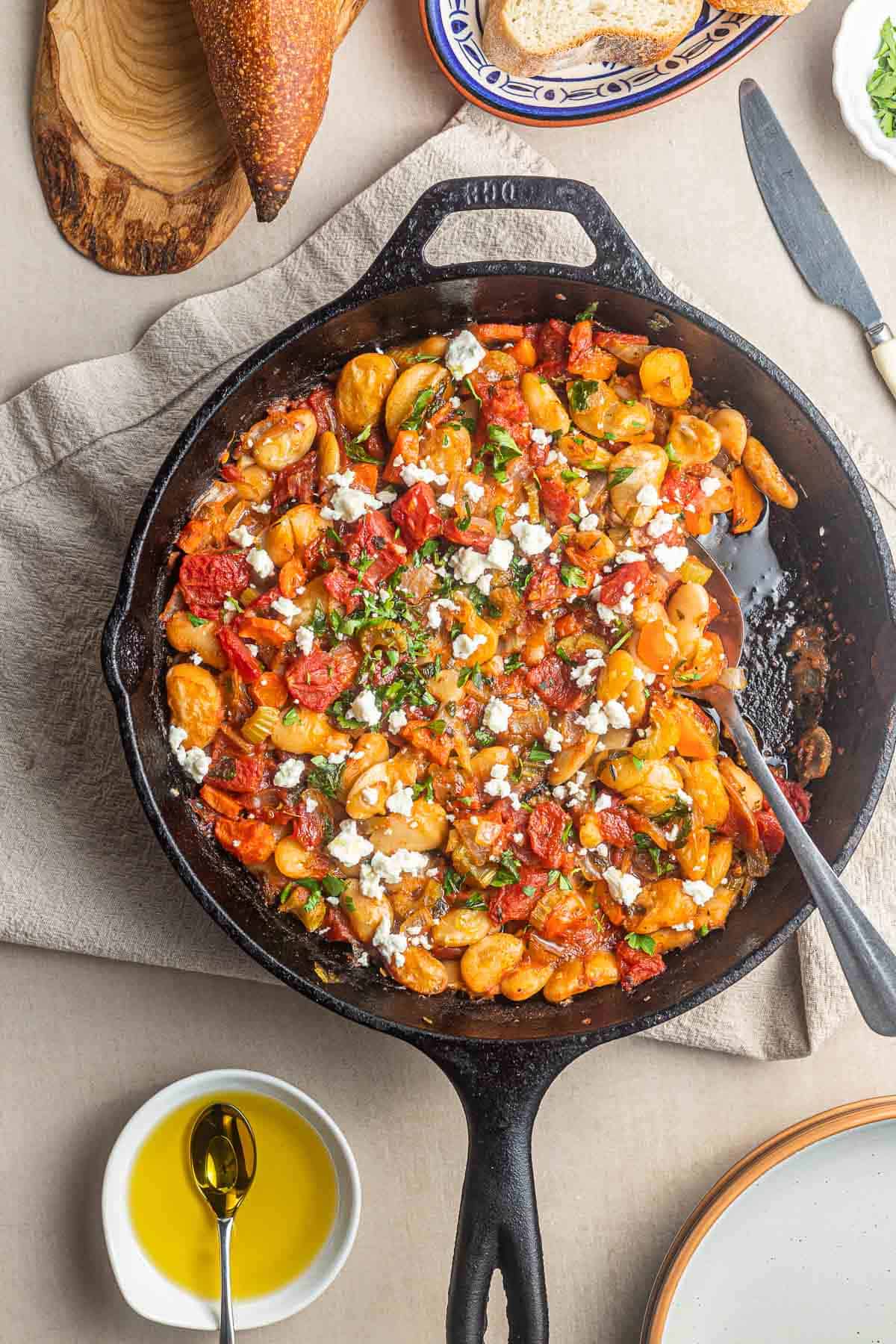 cooked gigantes plaki in a cast iron skillet with a spoon next to a bowl of olive oil with a spoon.