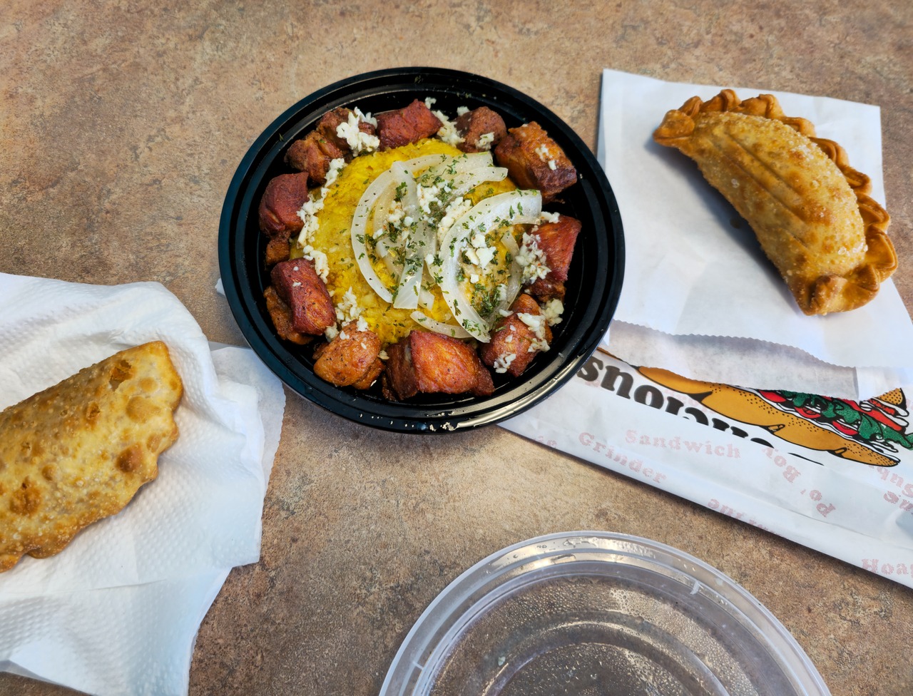 A black plastic container of mofongo in the middle of a table, surrounded by two empanadas.