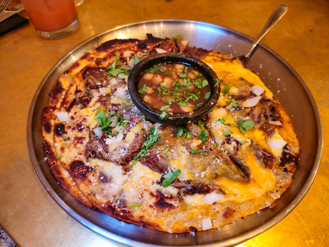 A tortilla "pizza" topped with shredded beef, cheese, onions and other ingredients, served with a side of consomme.