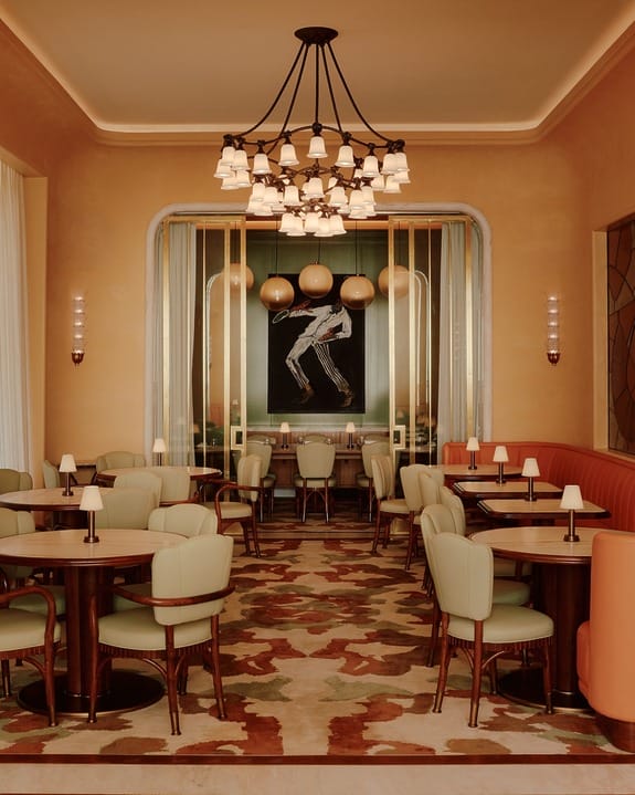 Elegant restaurant interior with soft lighting, round tables, cushioned chairs, a central chandelier, and art on the walls