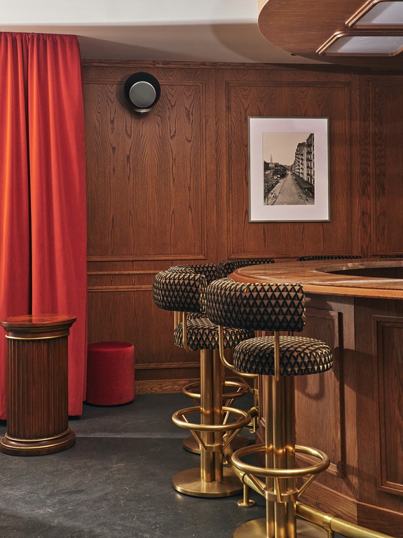 Stylish bar interior with wooden paneling, gold barstools, a red curtain, and a framed black-and-white photo on the wall.