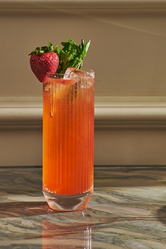 Tall glass of pink cocktail garnished with fresh herbs and a strawberry on marble table, surrounded by pastries and a wine glass.