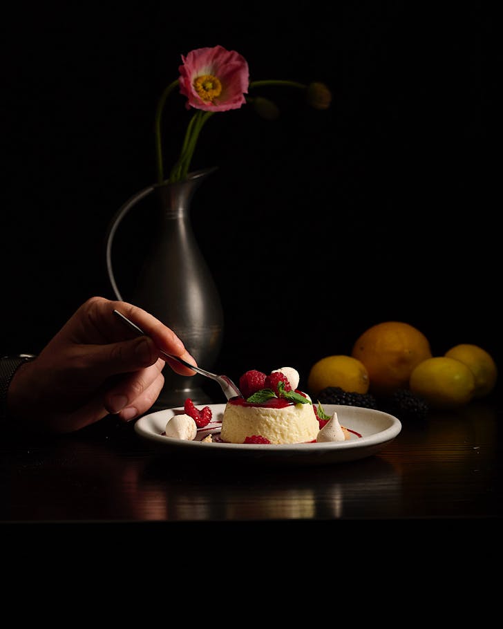 A plate of panna cotta topped with raspberries. A vase with a single pink flower sits in the background.