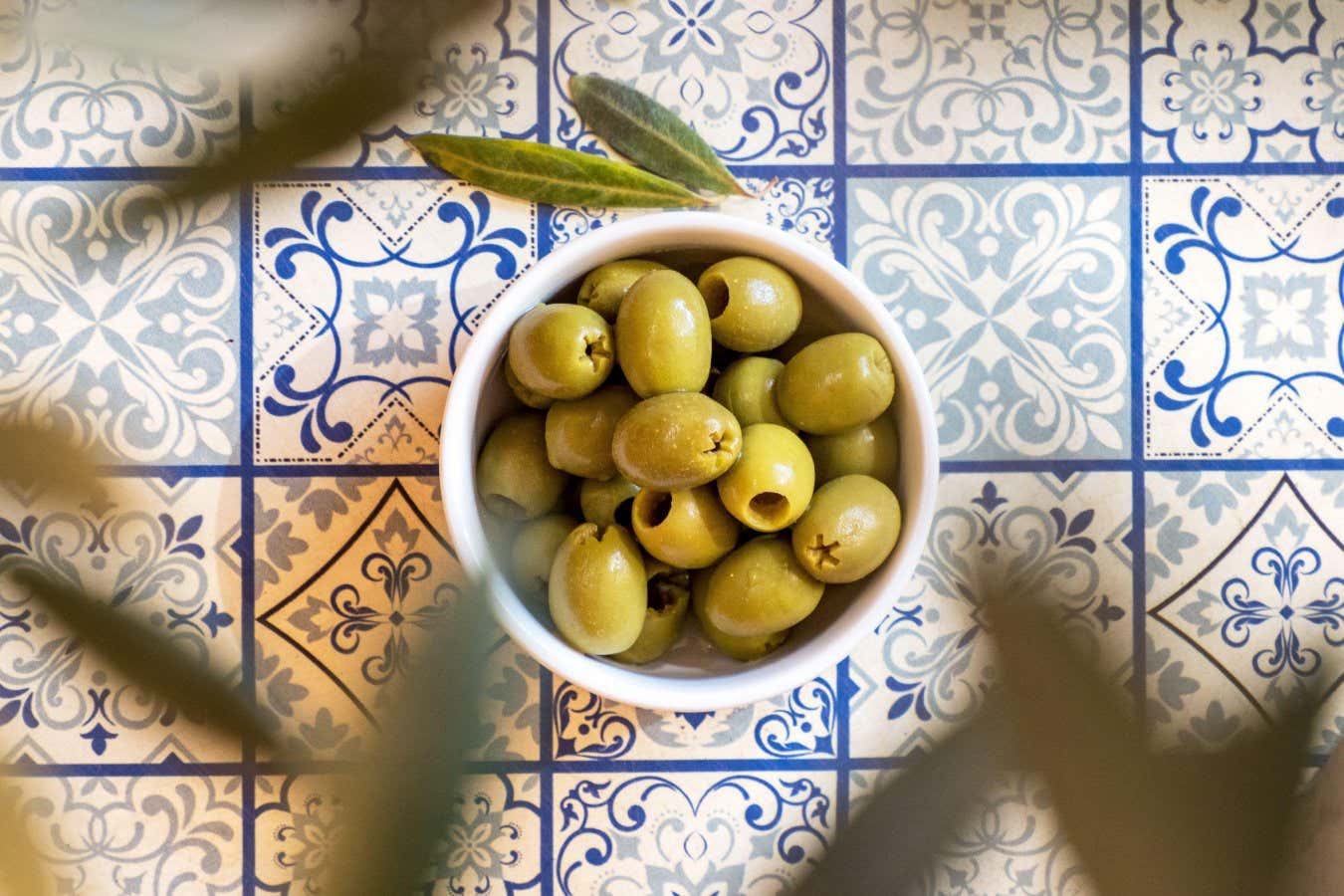 2GF0NFM Small white bowl with a green olives snack on a table with traditional Mediterranean blue pattern tiles under an olive tree