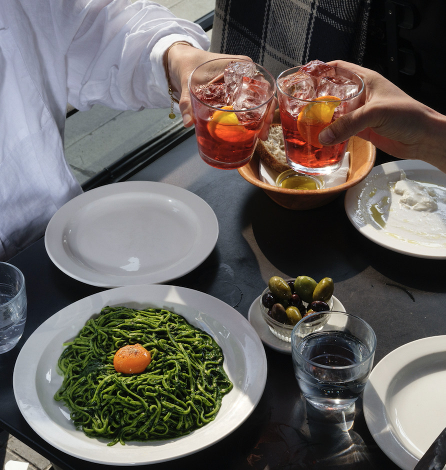 two negronis being cheersed over a plate of pasta and a small bowl of olives