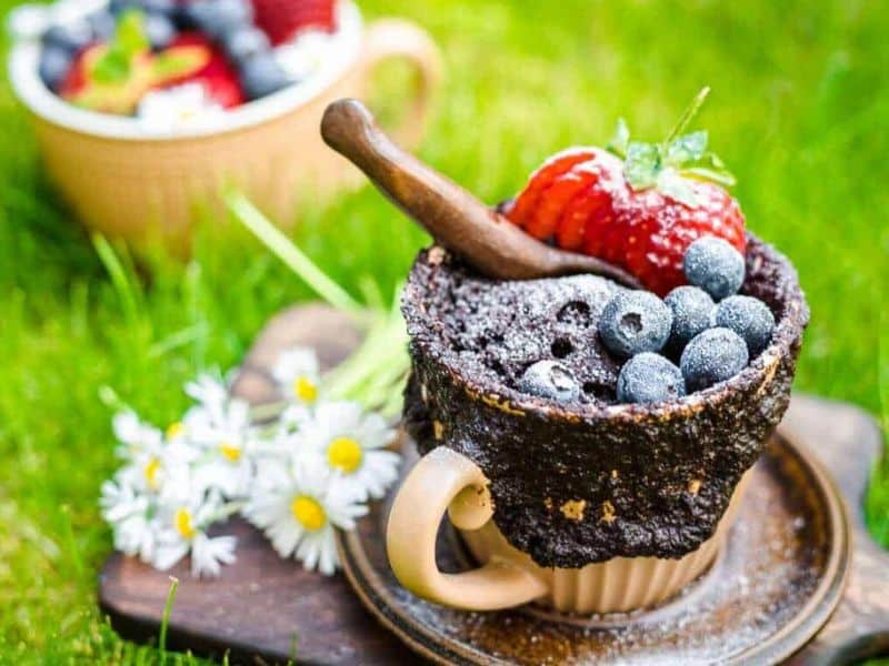 A cup-shaped chocolate dessert topped with blueberries, a sliced strawberry, powdered sugar, and a small wooden spoon, with daisies and another dessert in the background.