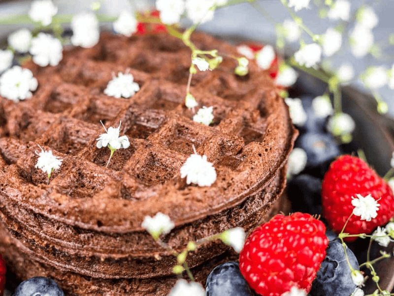 A stack of chocolate waffles garnished with small white flowers, surrounded by fresh raspberries and blueberries.