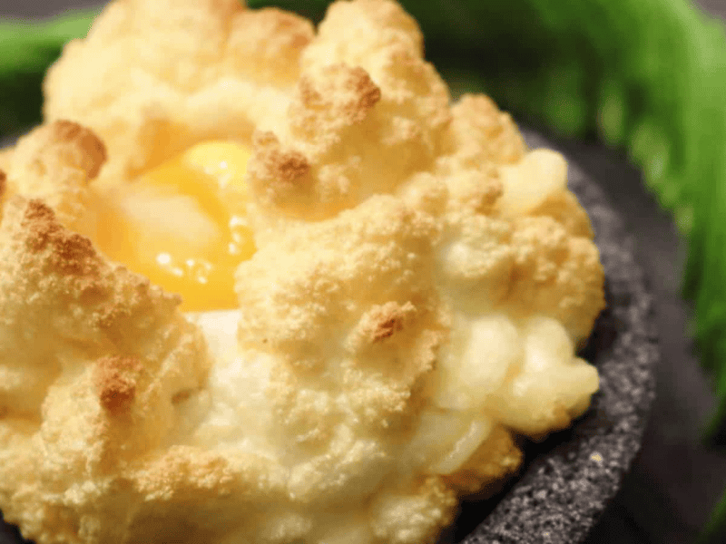 A close-up of a baked egg with fluffy, golden-brown whipped egg whites, resembling a cloud, served on a dark plate.