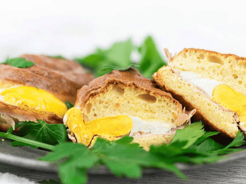 A close-up of a sandwich made with eggs, prosciutto, and cheese on a golden bun, served on a plate garnished with fresh parsley.